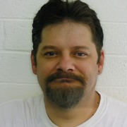 Man with dark hair, mustache, and goatee and a wry smile
